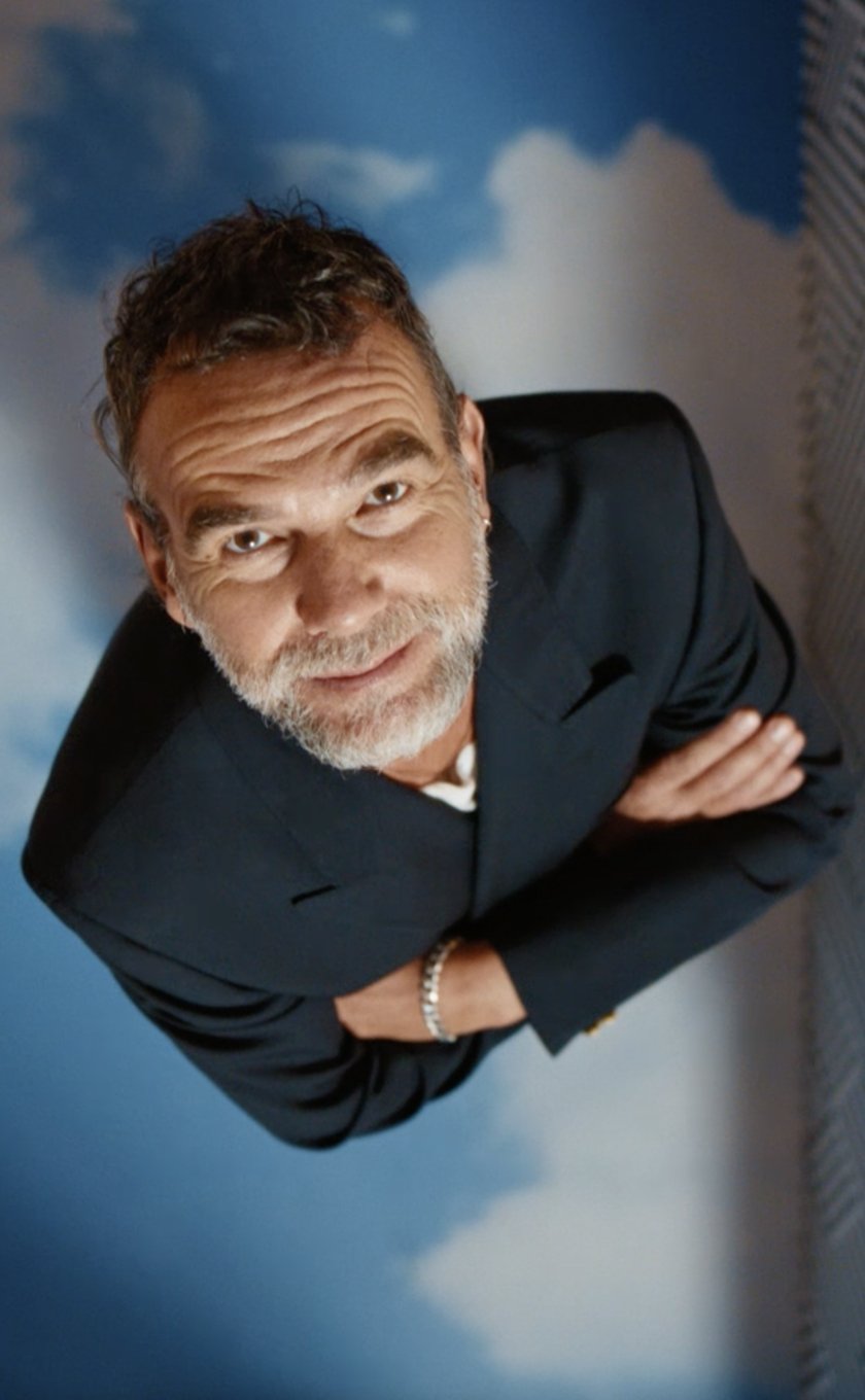 Stefano Seletti yukarıya bakıyor. Arka planda açık mavi gökyüzü, zikzak desenler ve bazı tasarım parçaları yer alıyor.